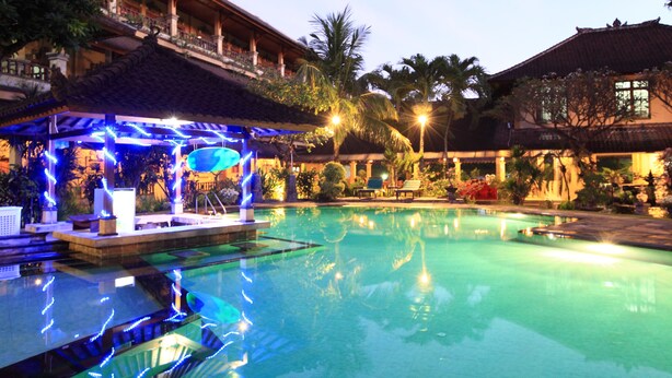 A swimming pool with a lounge area, palm trees, and a building with a tiled roof. - Balisandy Resort (Kuta)