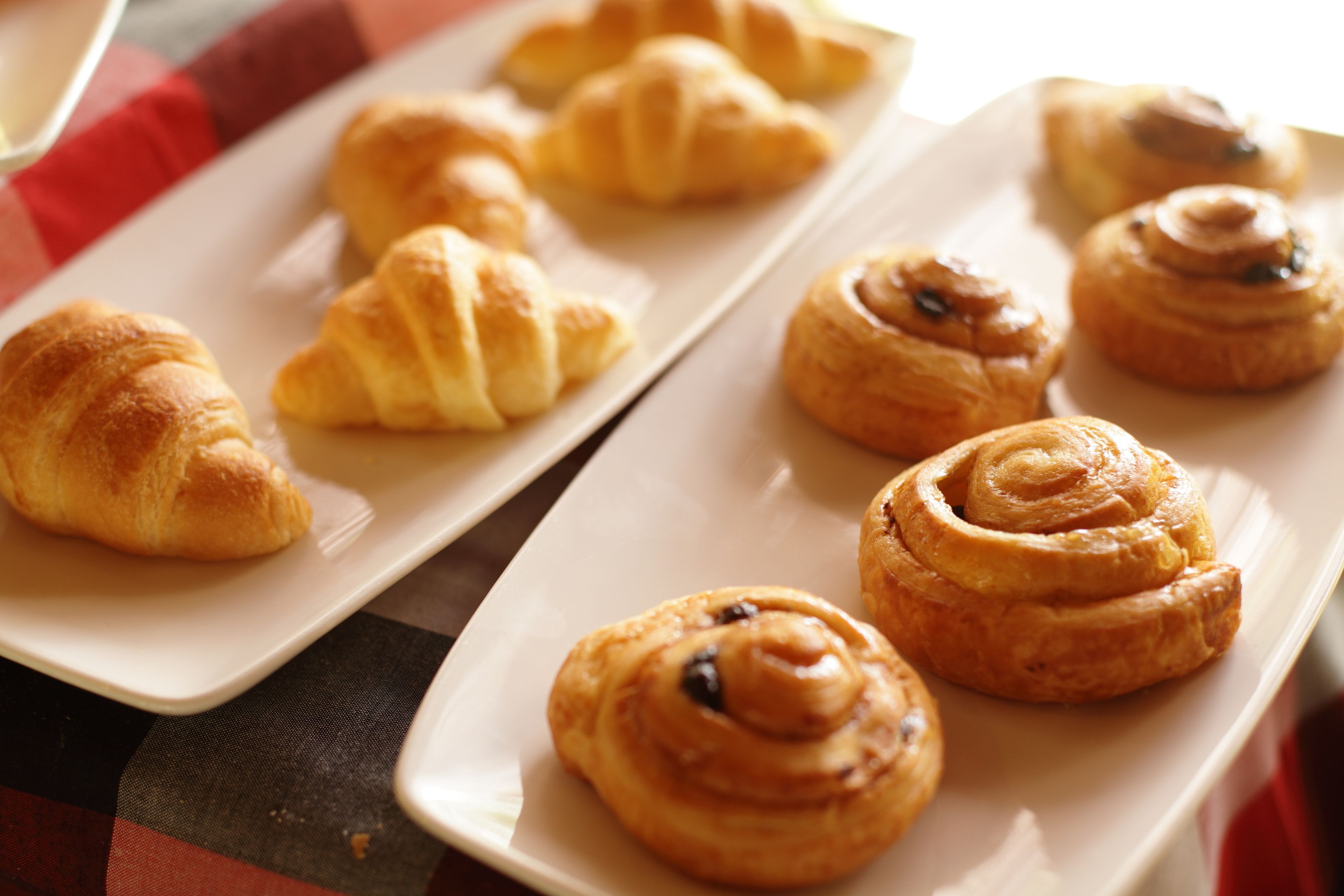 Une assiette remplie de diverses pâtisseries, notamment des croissants et des viennoiseries à la cannelle, est posée sur une table recouverte d’une nappe à carreaux.