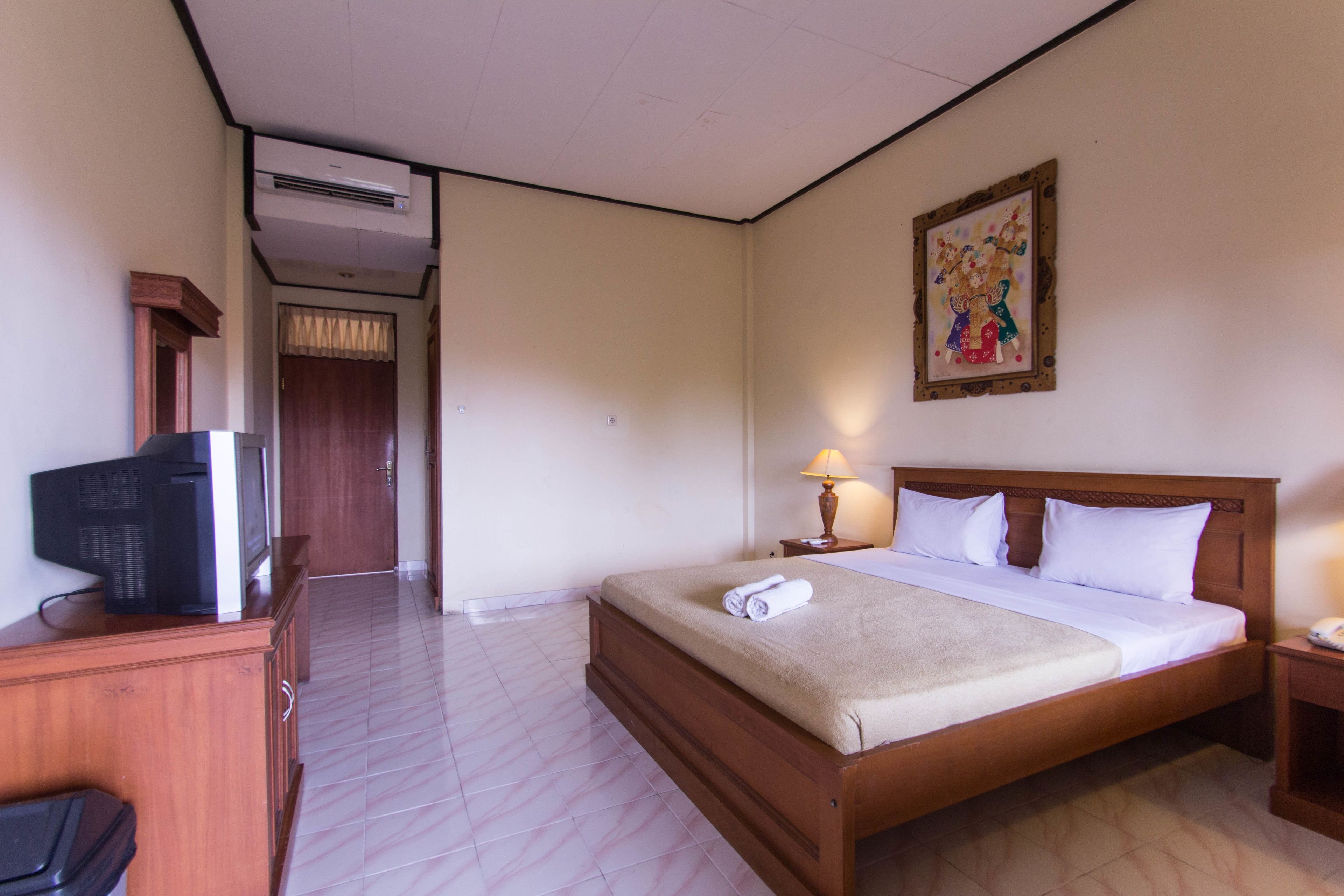 A hotel room with a wooden bed, a television on a wooden cabinet, a wall-mounted air conditioner, a bedside lamp, and a painting on the wall.