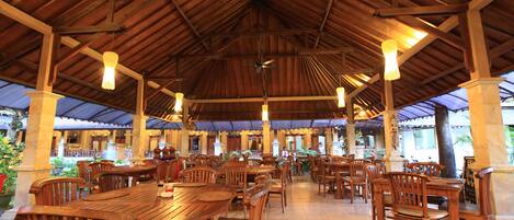 A spacious covered dining area with wooden tables and chairs, a high ceiling with exposed beams, and hanging pendant lights.