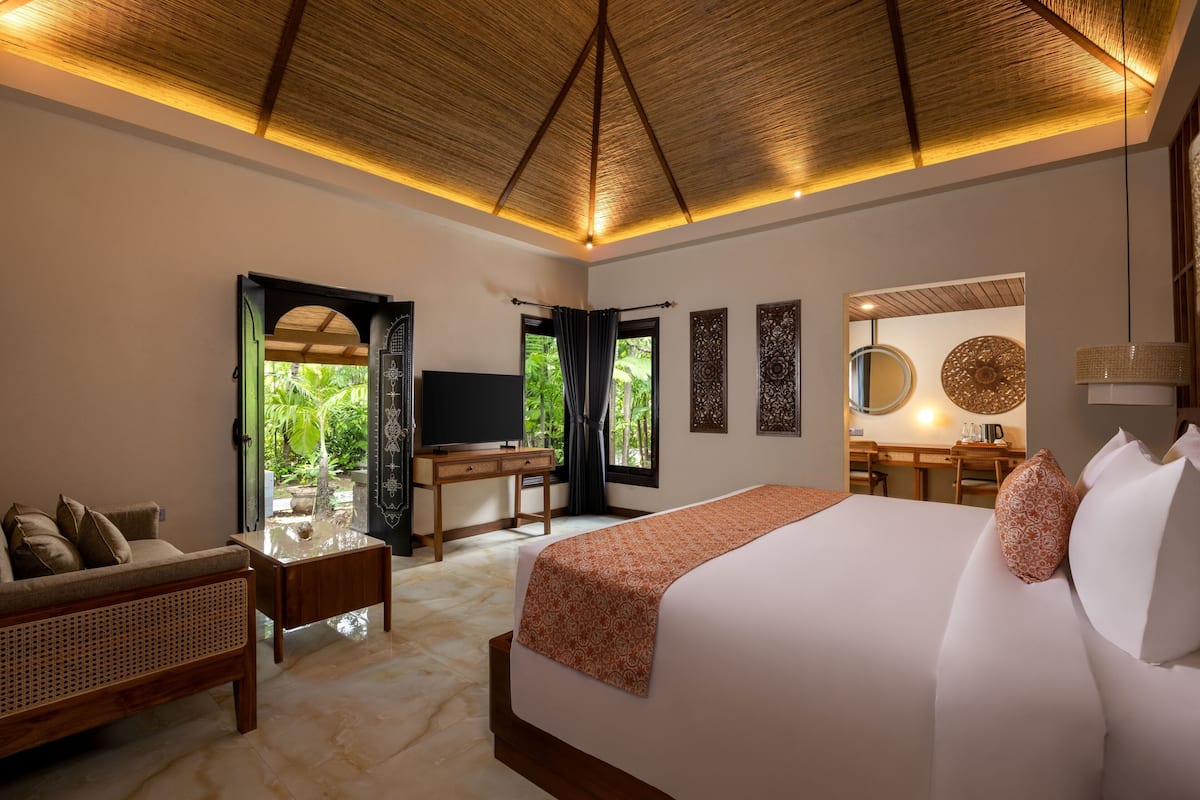 A well-lit bedroom with a large bed, a wooden desk, a television, and a view of greenery outside.
