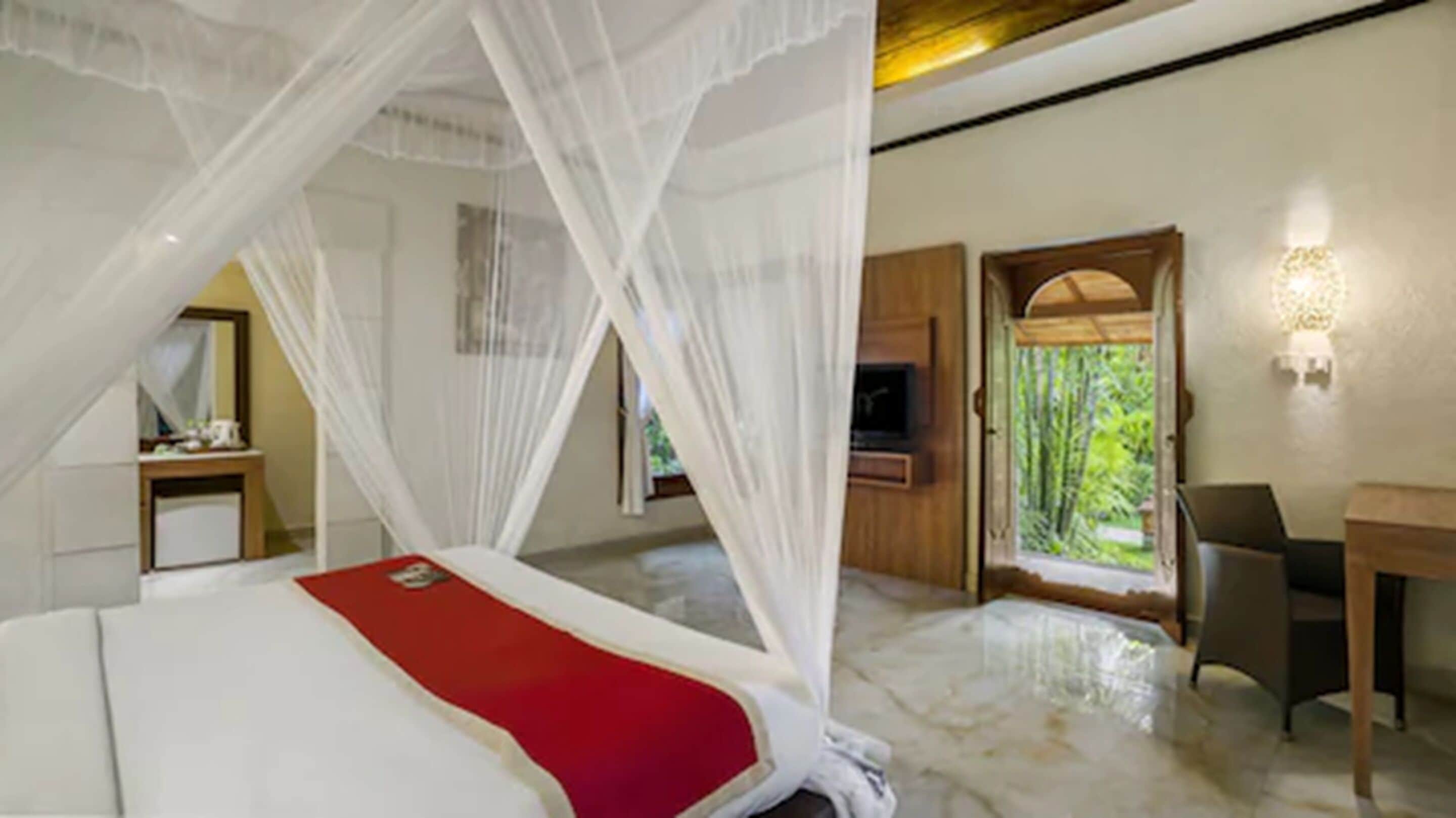 A bedroom with a bed, a TV, a desk, and a window with a view of greenery.