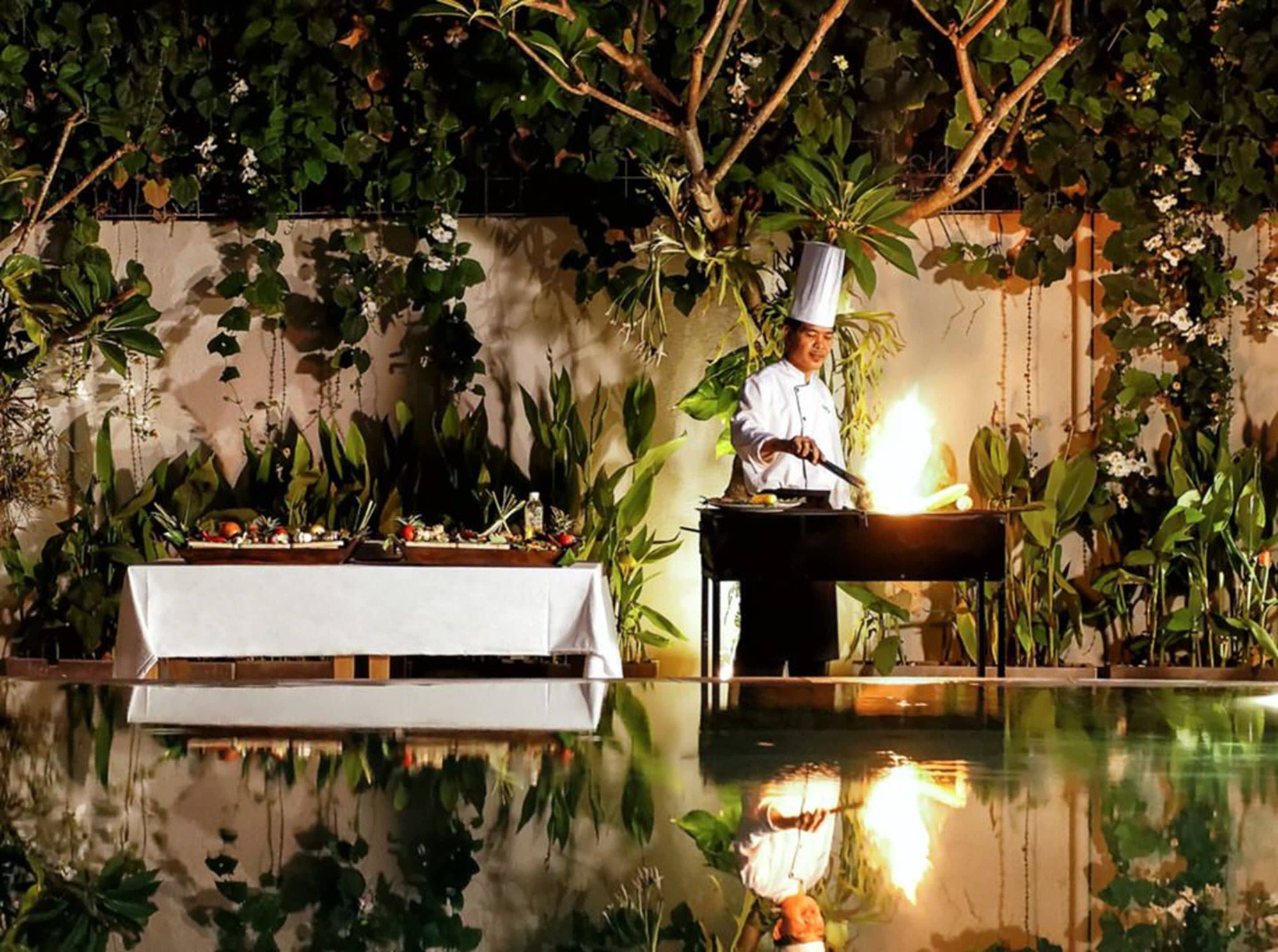 A chef cooking outdoors with a table set for a meal, surrounded by lush greenery and a reflective pool.