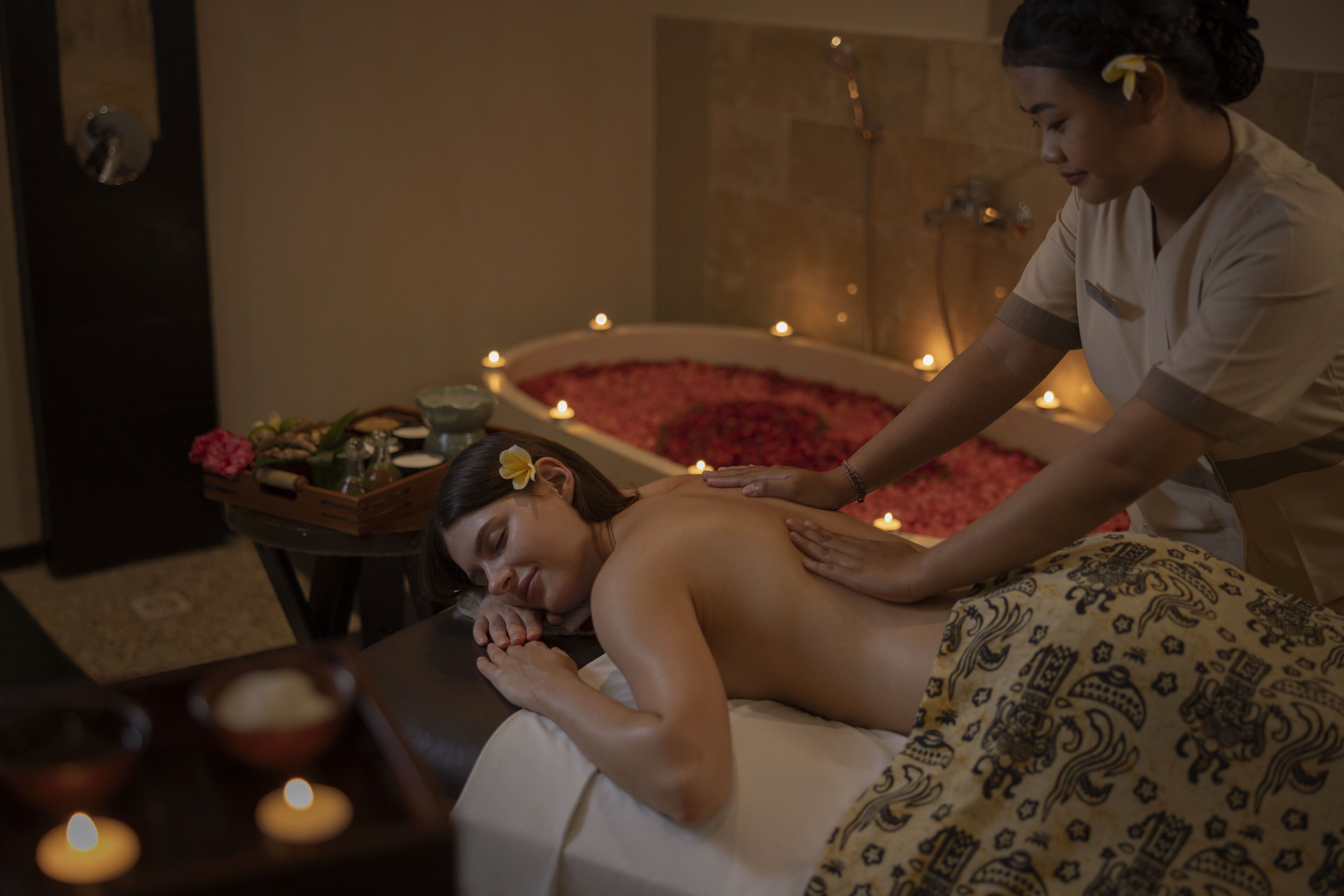 Eine Person erhält eine Massage in einem Raum mit einer Badewanne, die mit Rosenblättern, Kerzen und einer Ablage mit Blumen gefüllt ist.