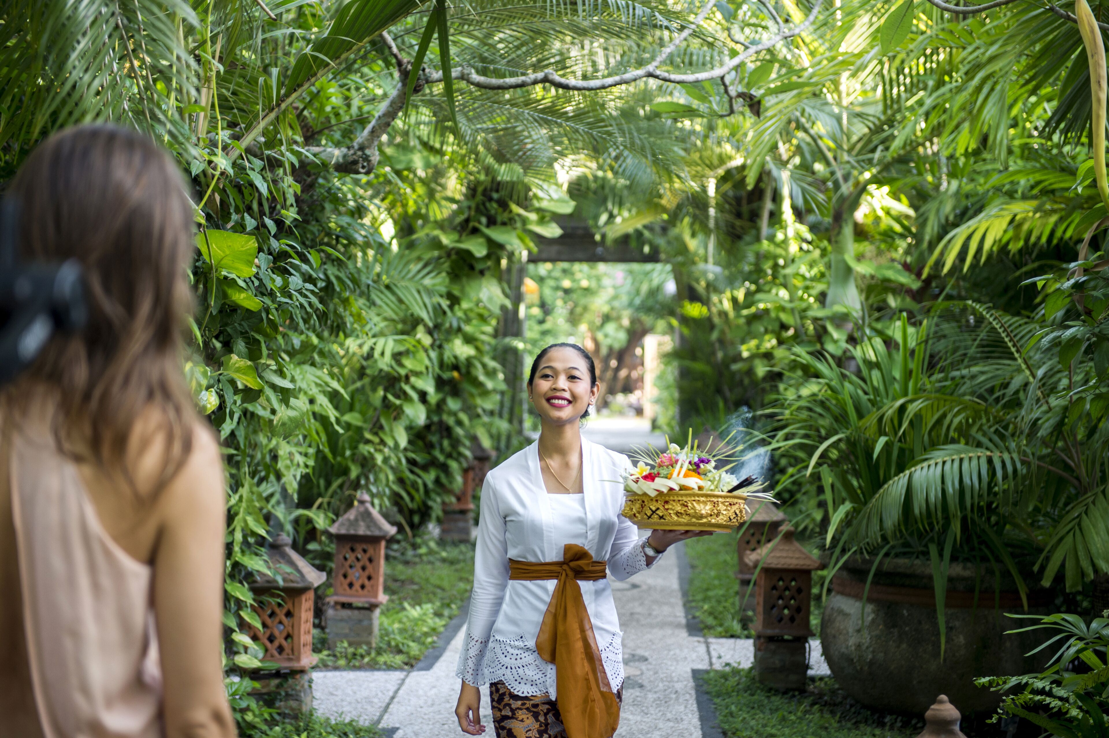 A person holding a basket with flowers, standing in a lush garden with a pathway and lanterns.