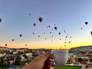 Une main tenant une tasse, avec en arrière-plan une vue sur une ville et de nombreux ballons à air chaud dans le ciel.