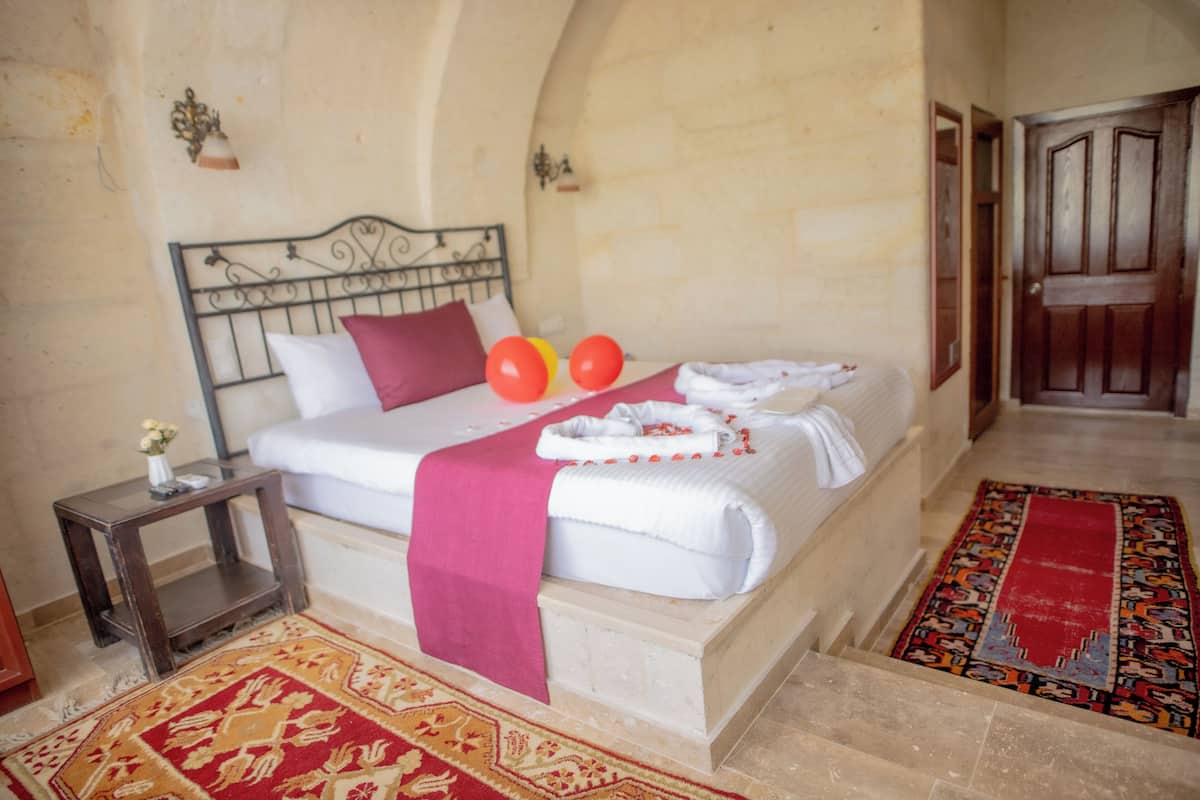A bedroom with a bed, pillows, a bedside table, a nightstand, a red and beige patterned rug, and a wooden door.