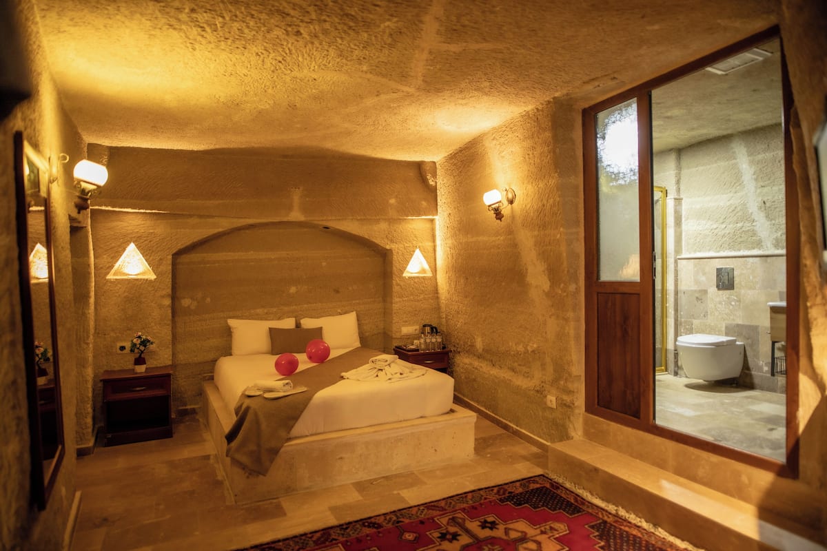 A bedroom with a large bed, a nightstand, a bedside table, a window, and a view of the bathroom.