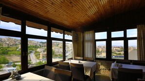 Un comedor con mesas y sillas, ventanas amplias con cortinas y vistas a un paisaje urbano.