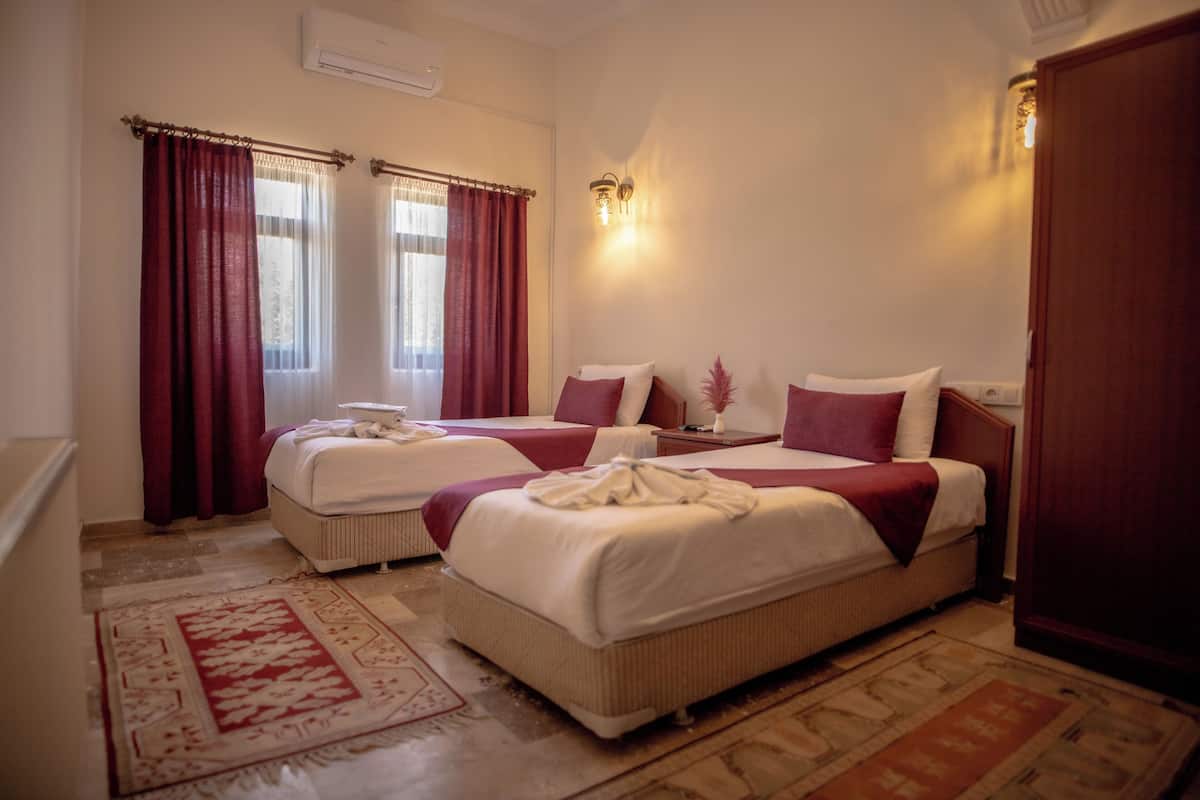 A hotel room with two beds, a large window with curtains, a wall-mounted light fixture, and a wooden door.