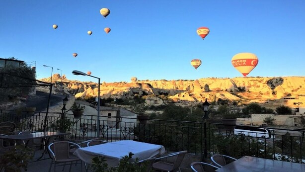 Una terraza con mesas y sillas, con vistas a un paisaje rocoso y globos aerostáticos al fondo.