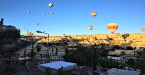 A terrace with tables and chairs, overlooking a rocky landscape with hot air balloons in the background. - Guven Cave Hotel (Nevsehir)