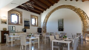 Un comedor con mesas blancas, sillas de mimbre, arcos de piedra y techo de madera rústica.