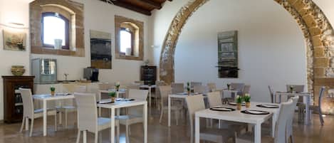 Un comedor con mesas blancas, sillas de mimbre, arcos de piedra y techo de madera rústica.