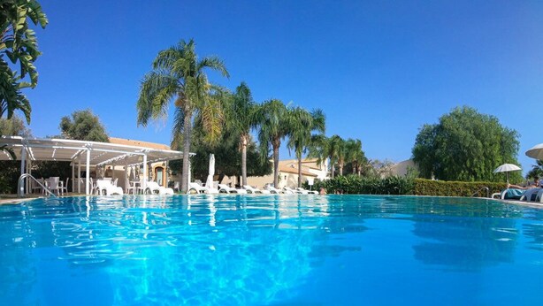 Una piscina azul cristalina con sillas de descanso y sombrillas, rodeada de palmeras y una zona de estar cubierta.