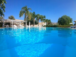 A clear blue pool with lounge chairs and umbrellas, surrounded by palm trees and a covered seating area. - Life Hotels Kalaonda Resort (Syracuse)