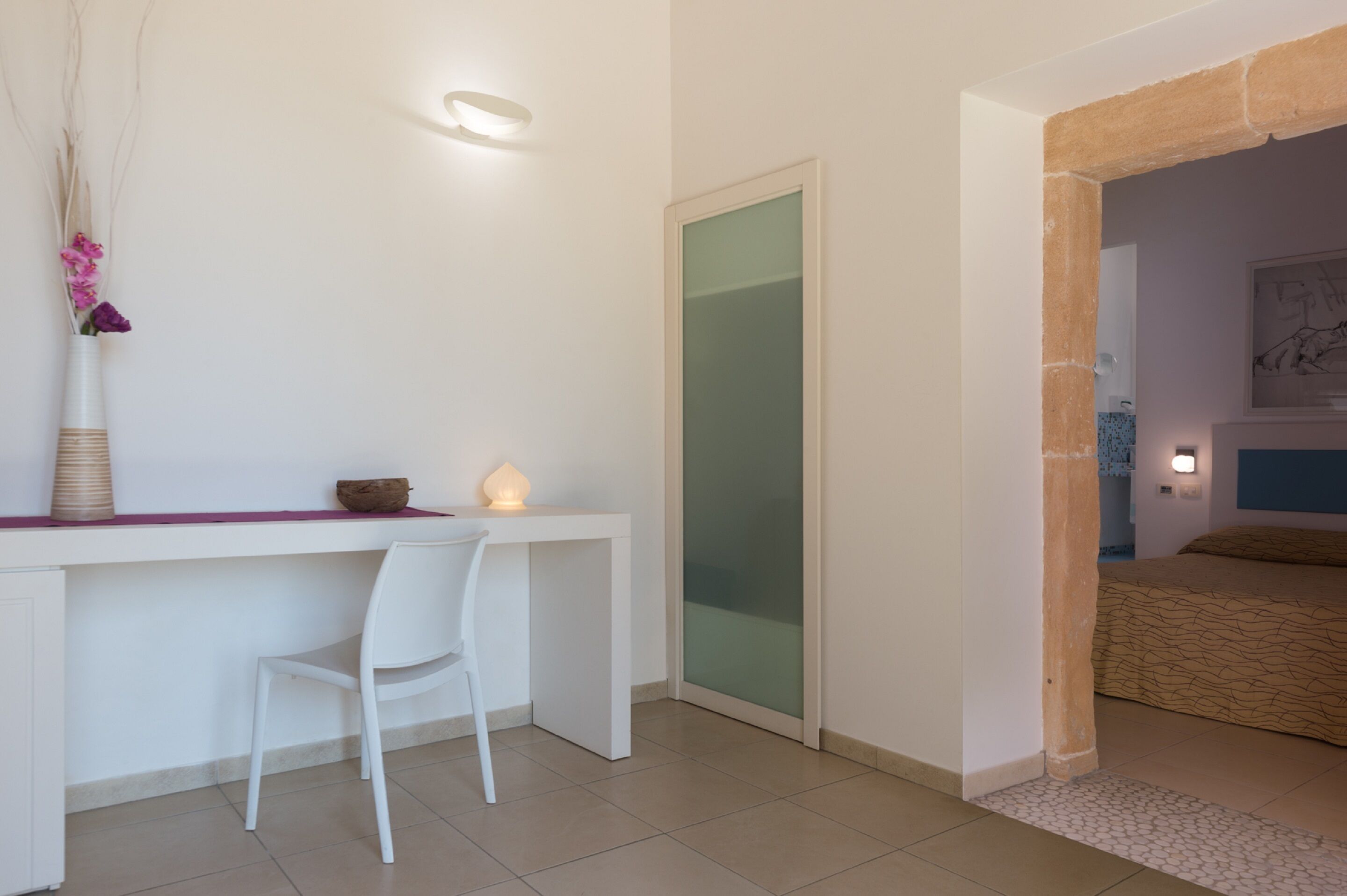 Une chambre moderne avec un bureau blanc, une chaise blanche, une porte en verre et un lit recouvert d’une couvre-lit à motifs.