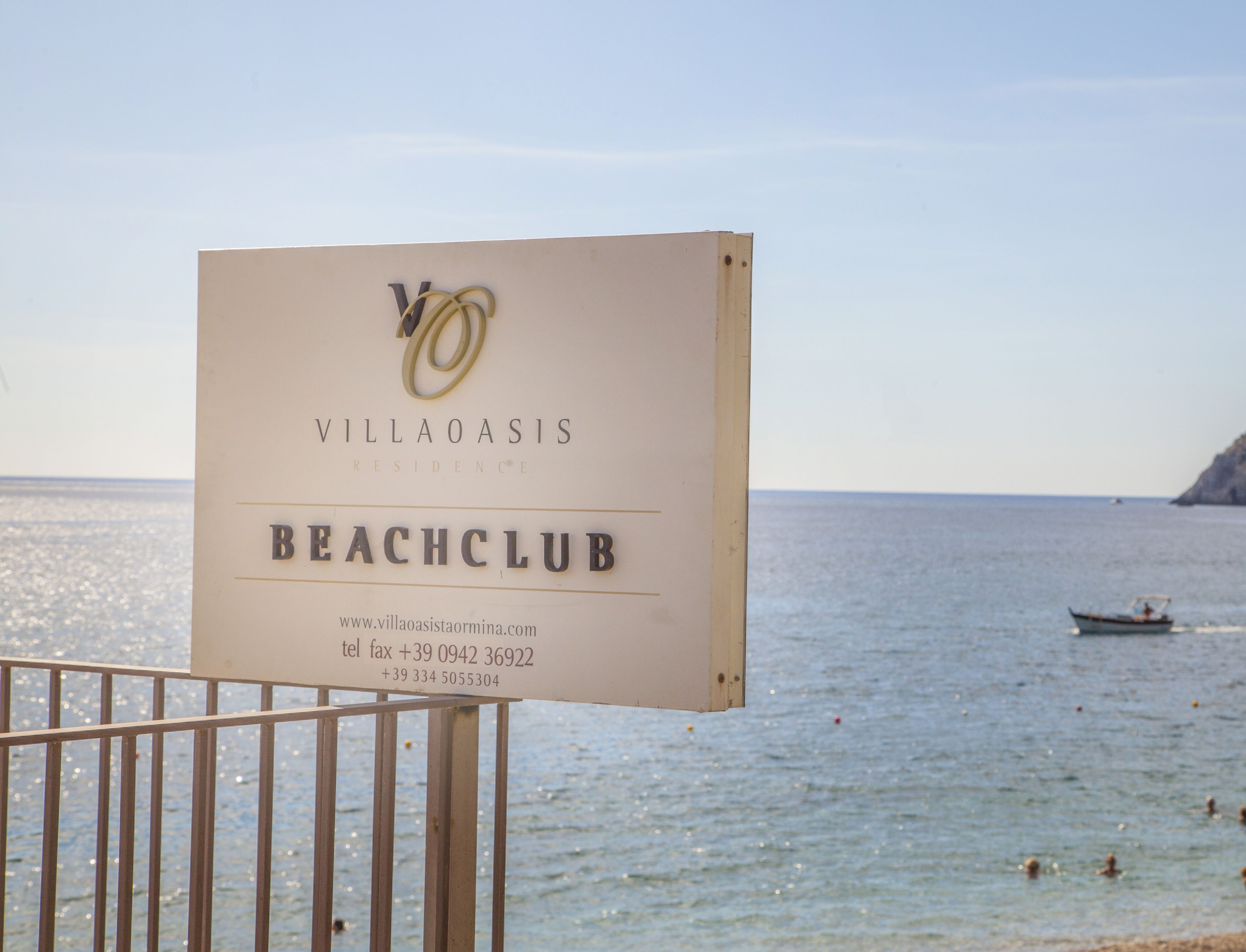 Schild für den Villa Oasis Beach Club mit Kontaktinformationen und Blick auf das Meer.