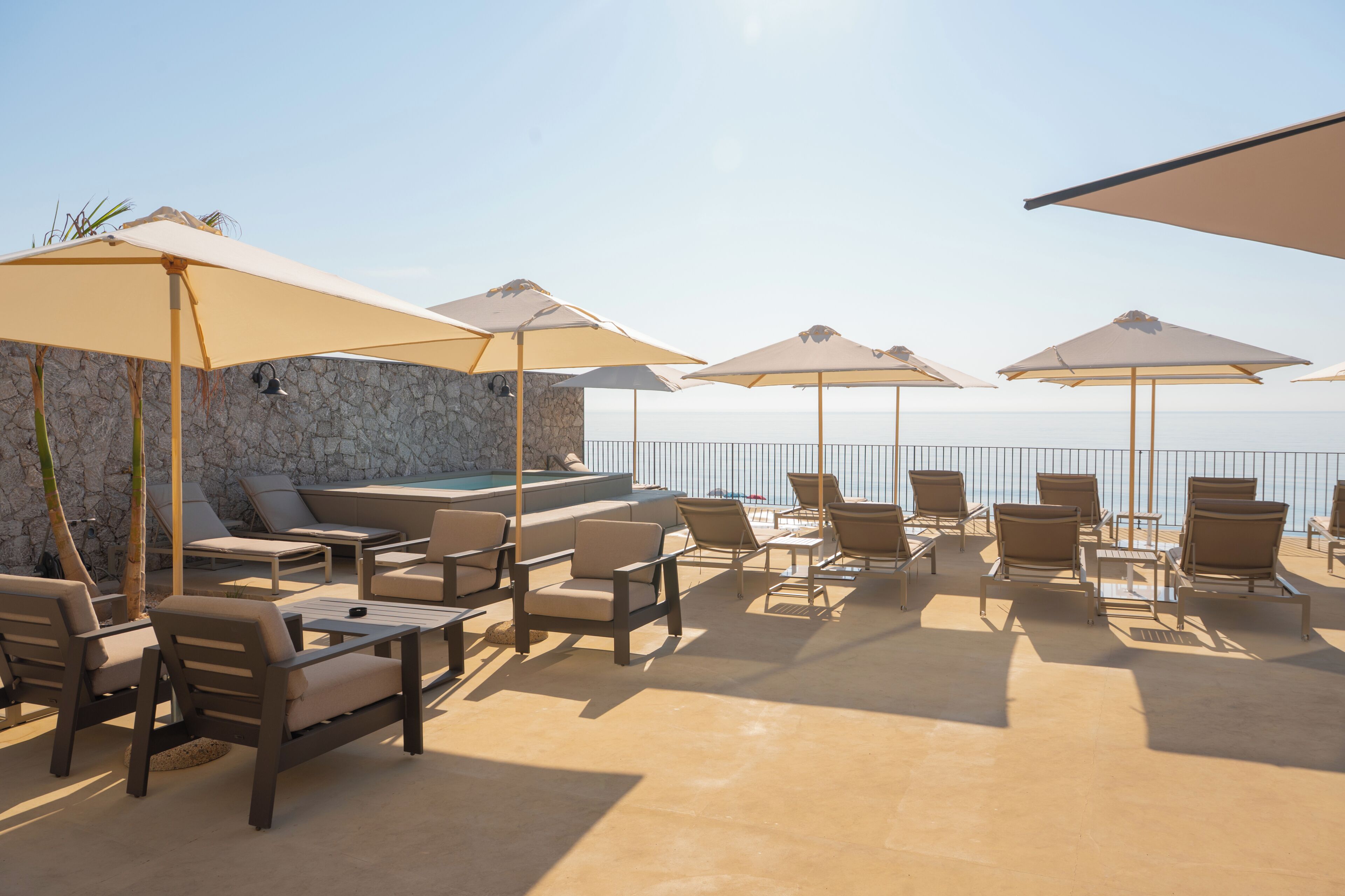 Área externa com assentos de lounge, cadeiras e guarda-sóis, uma parede de pedra e vista para o mar.