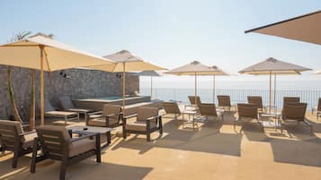 Área externa com assentos de lounge, cadeiras e guarda-sóis, uma parede de pedra e vista para o mar.