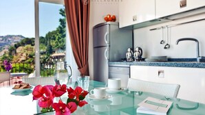 A modern kitchen with a glass table, white cabinets, and a view of a mountainous landscape. - Villa Oasis Residence (Taormina)