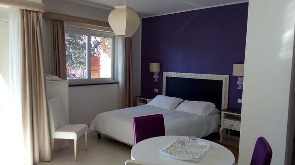 A hotel room with a bed, bedside tables, a chair, a small table, and a window with curtains. - Villa Oasis Residence (Taormina)