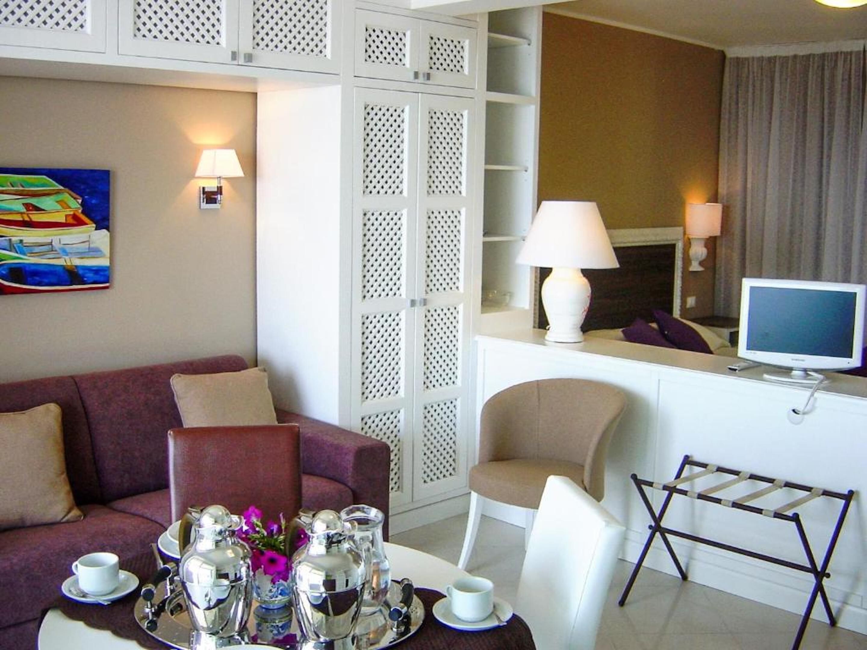 Una camera d'albergo con un divano, un tavolo da pranzo con un vassoio di posate e tazze, una TV e un quadro decorativo.