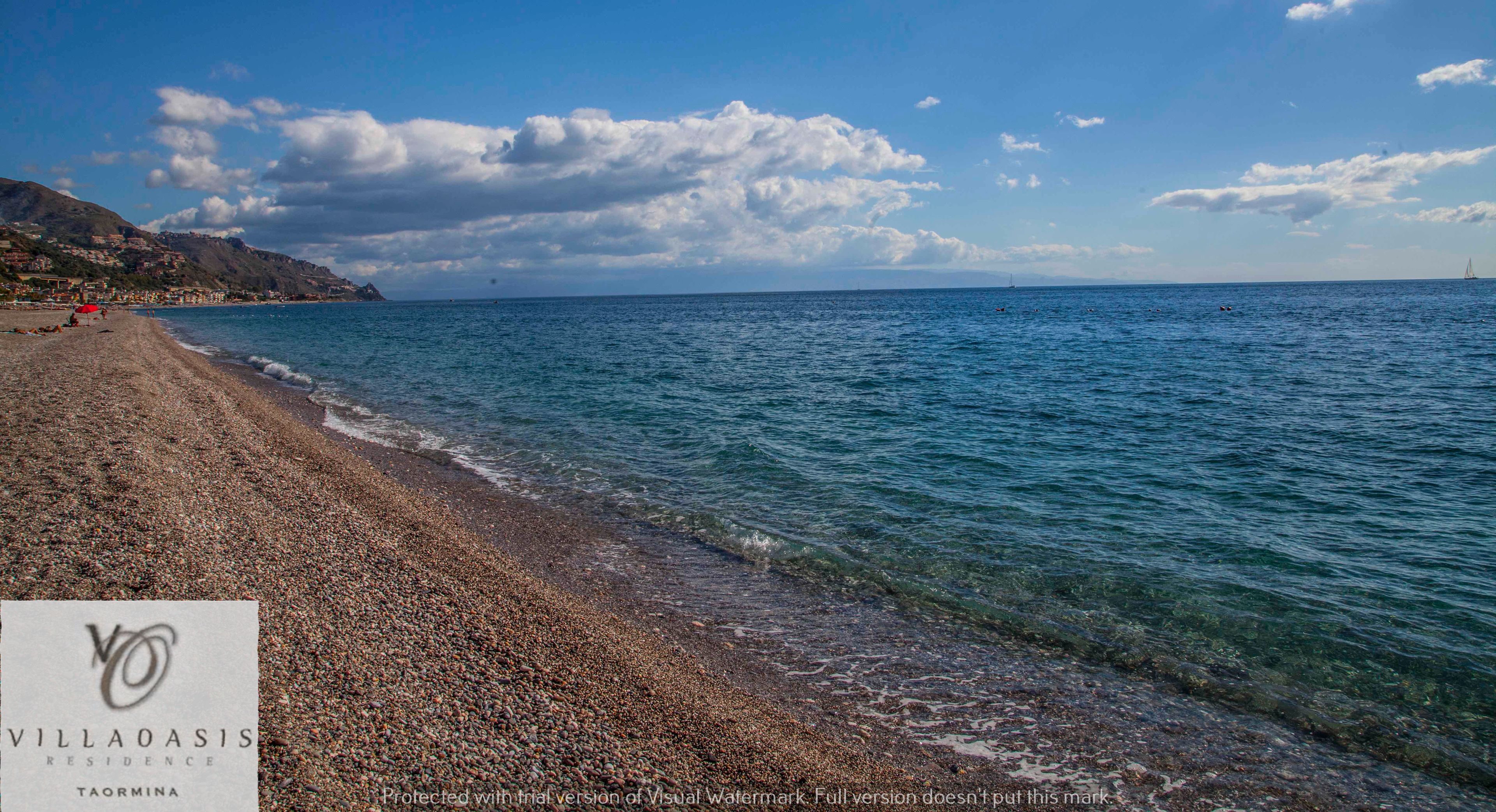 A beach with pebbles, a sign for Villa Oasis Residence Taormina, and a sailboat in the distance.