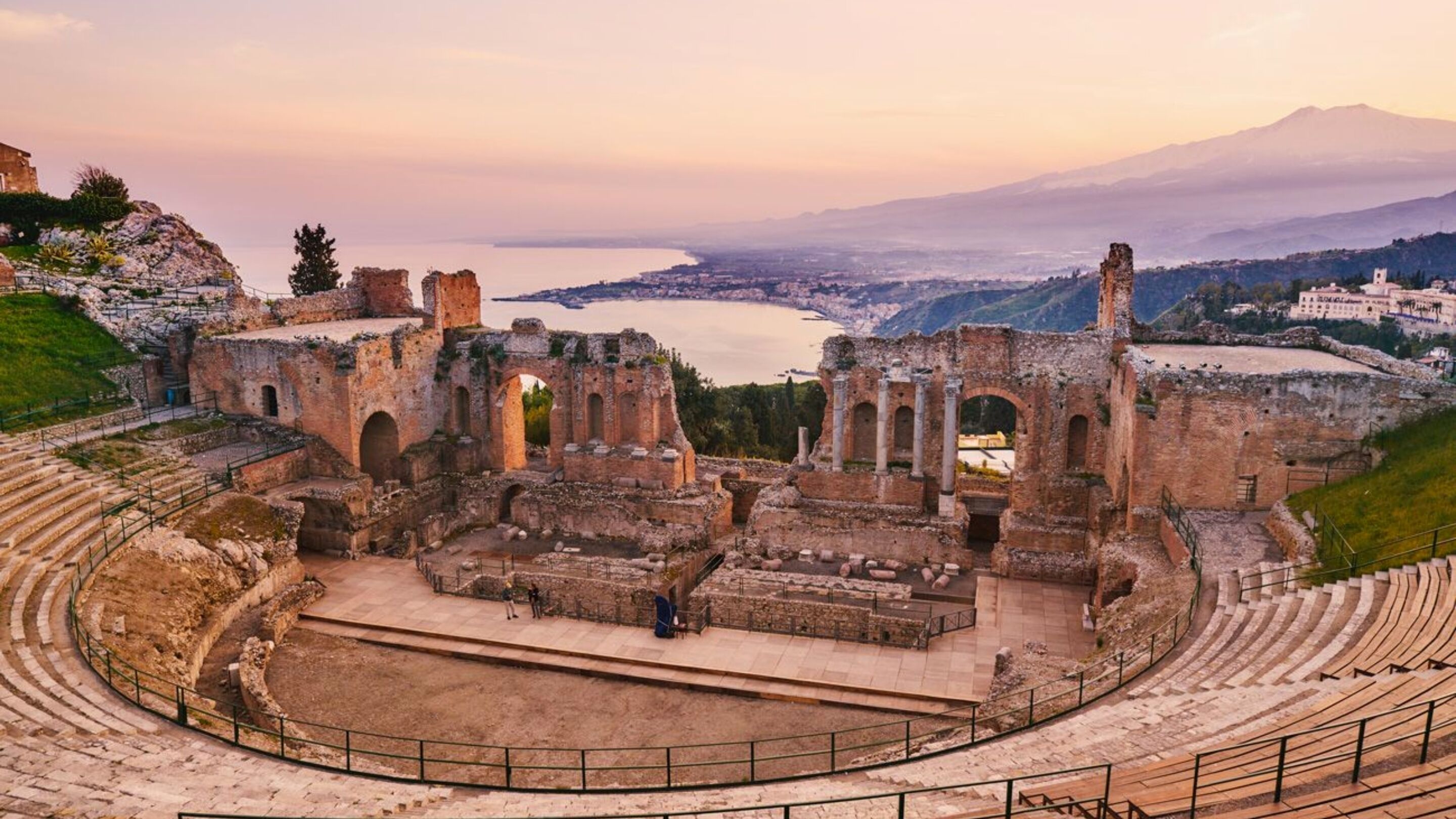 An ancient amphitheater with ruins and a view of a body of water and mountains in the background.