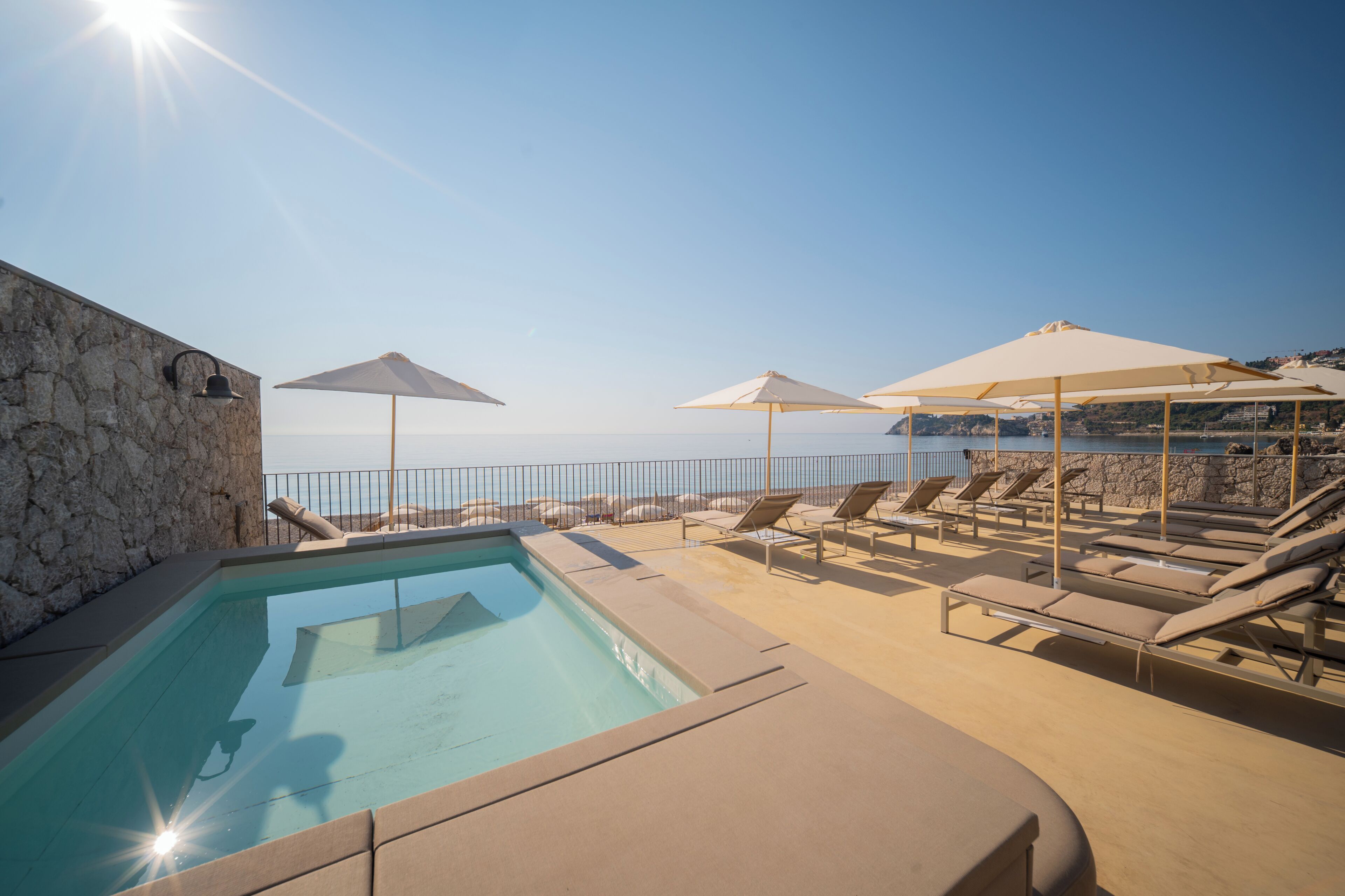 Zona de piscina con sillas de descanso y sombrillas, con vista al mar tranquilo.