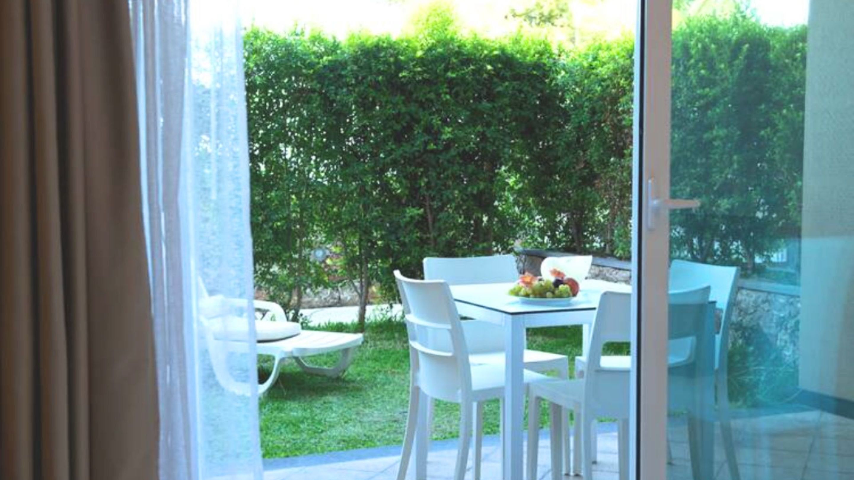 Un balcón con mesa y sillas, un ventanal de vidrio y vista a un seto verde.