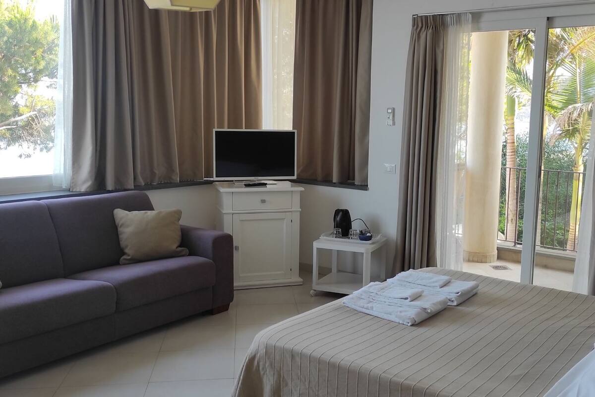 A hotel room with a bed, a sofa, a TV, a small table with a tray, and a balcony with curtains.