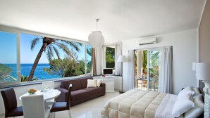 A modern hotel room with a large window offering a sea view, a cozy bed, a dining area, and a small table with a laptop. - Villa Oasis Residence (Taormina)