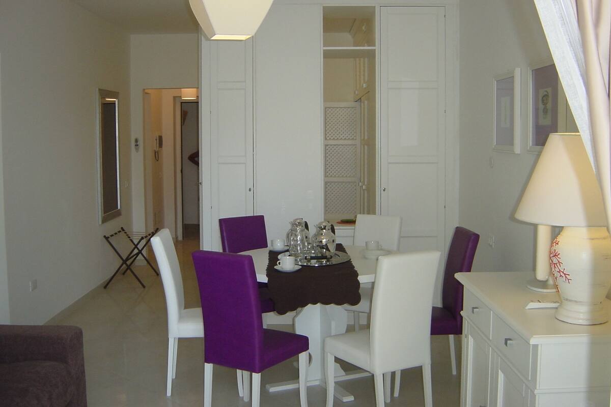 A dining area with a table set for four, surrounded by white chairs with purple upholstery. There is a white cabinet to the right and a white door in the background.