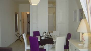 Un comedor con una mesa dispuesta para cuatro personas, rodeada de sillas blancas con tapicería morada. A la derecha hay un armario blanco y una puerta blanca al fondo.