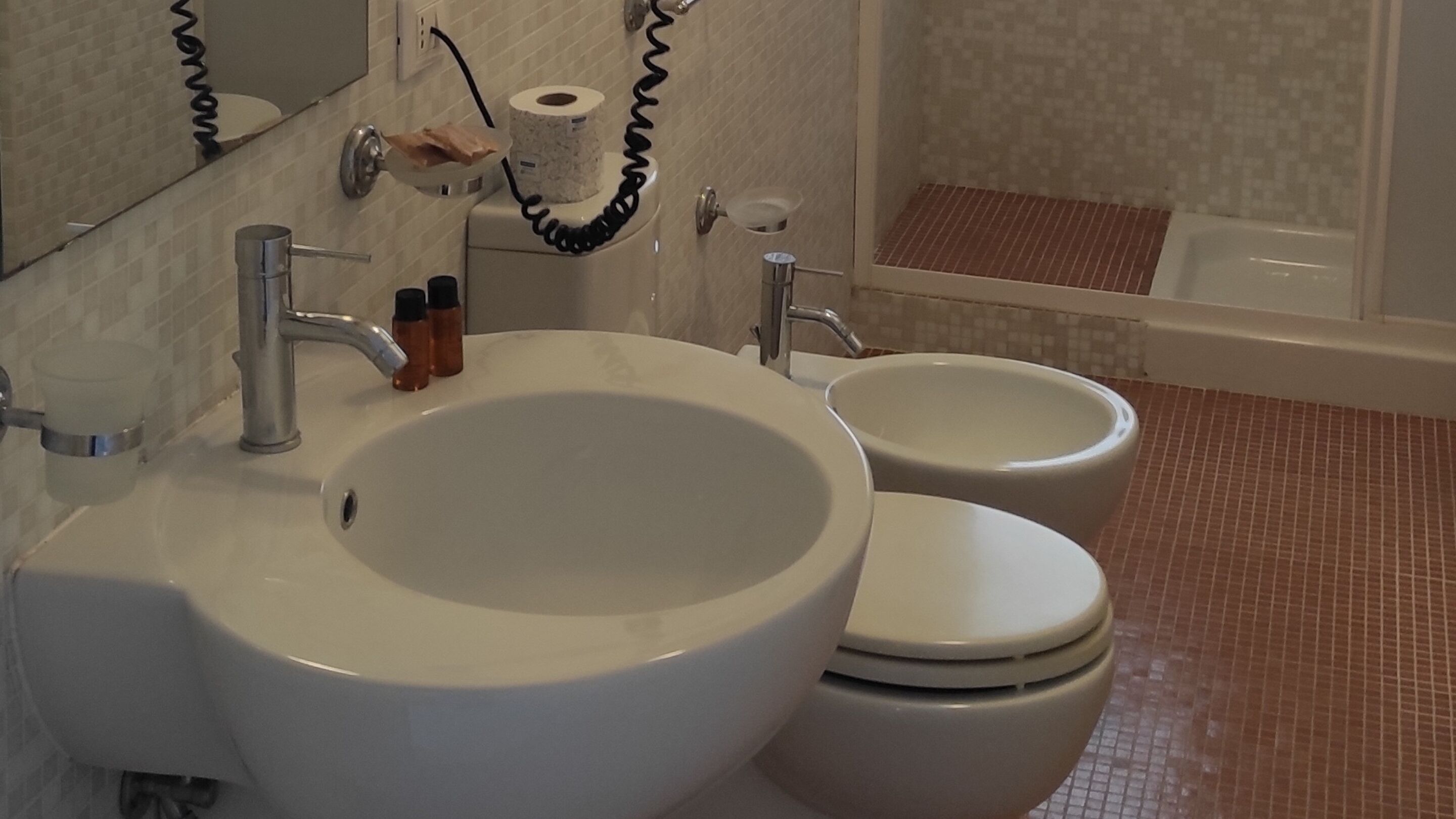 Un baño con lavamanos, inodoro y espejo. Hay un rollo de papel higiénico y un dispensador de jabón en la pared.