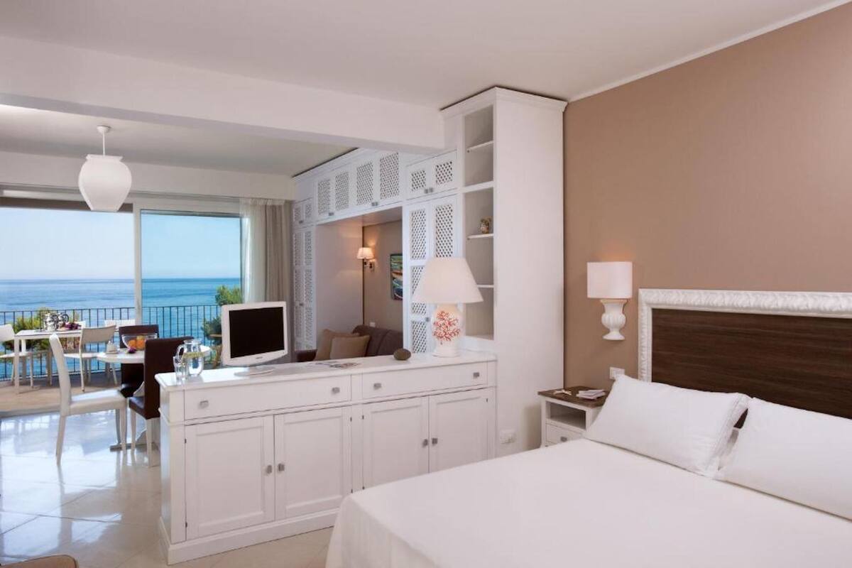 A hotel room with a bed, a desk with a computer, a TV, and a balcony with a view of the sea.