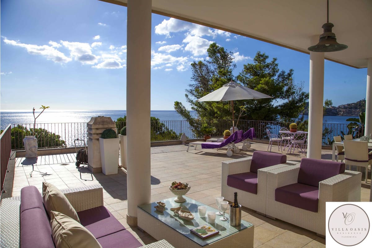 A covered outdoor seating area with wicker furniture, a table with a bottle and glasses, and a view of the sea.