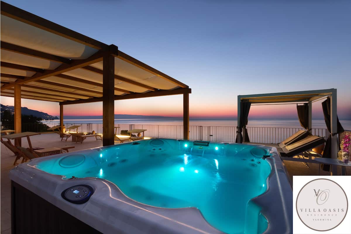 A hot tub with a control panel, surrounded by lounge chairs and tables, overlooking a sunset view.