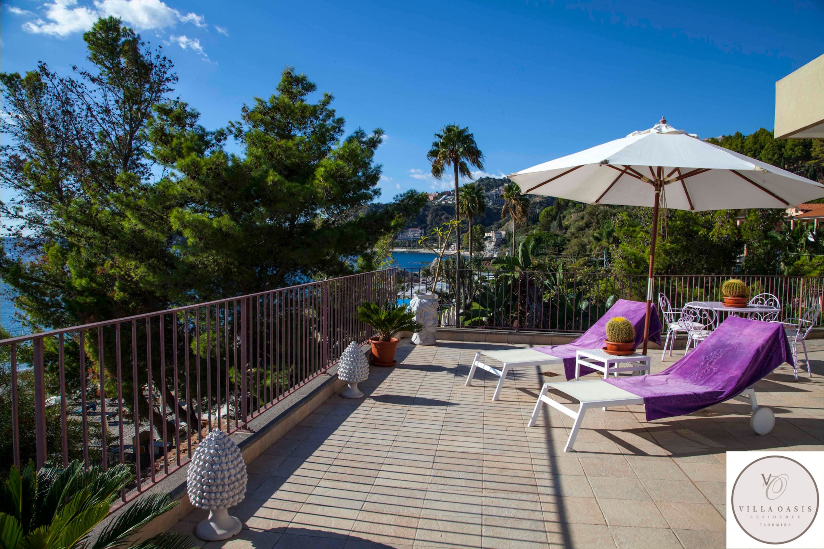 A terrace with white lounge chairs, a large white umbrella, and potted plants.