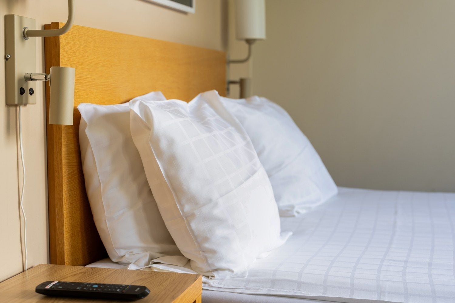 Een hotelkamer met een houten hoofdeinde, witte beddengoed en een afstandsbediening op een nachtkastje van hout.