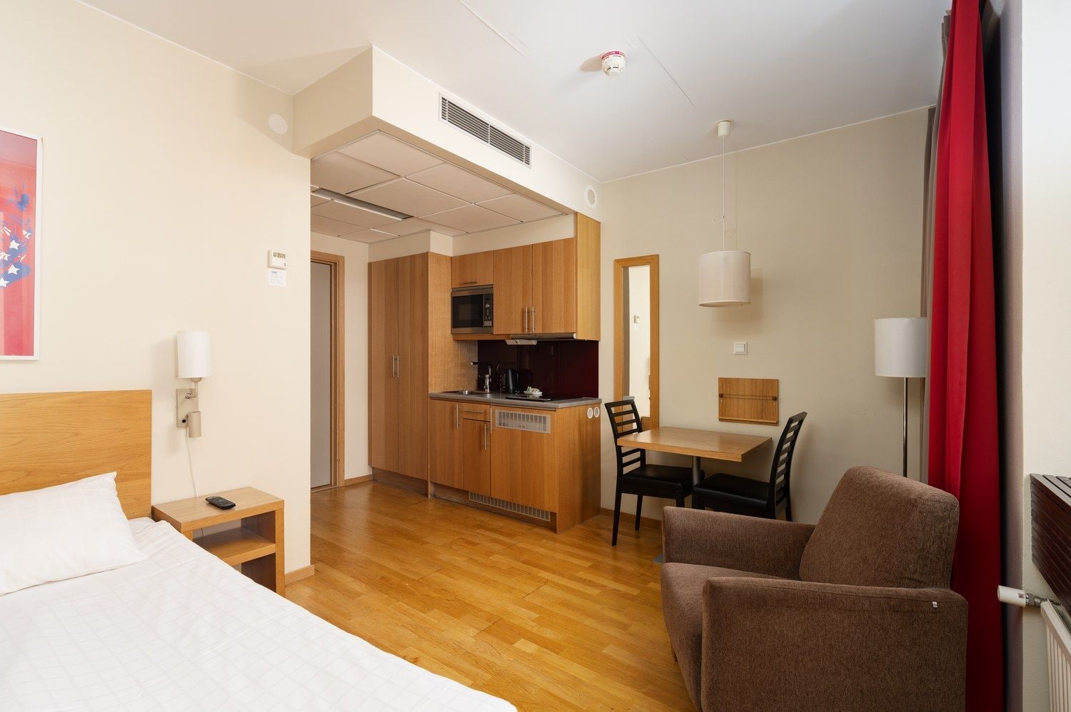 A hotel room with a bed, a desk, a chair, a kitchenette, and a bathroom.