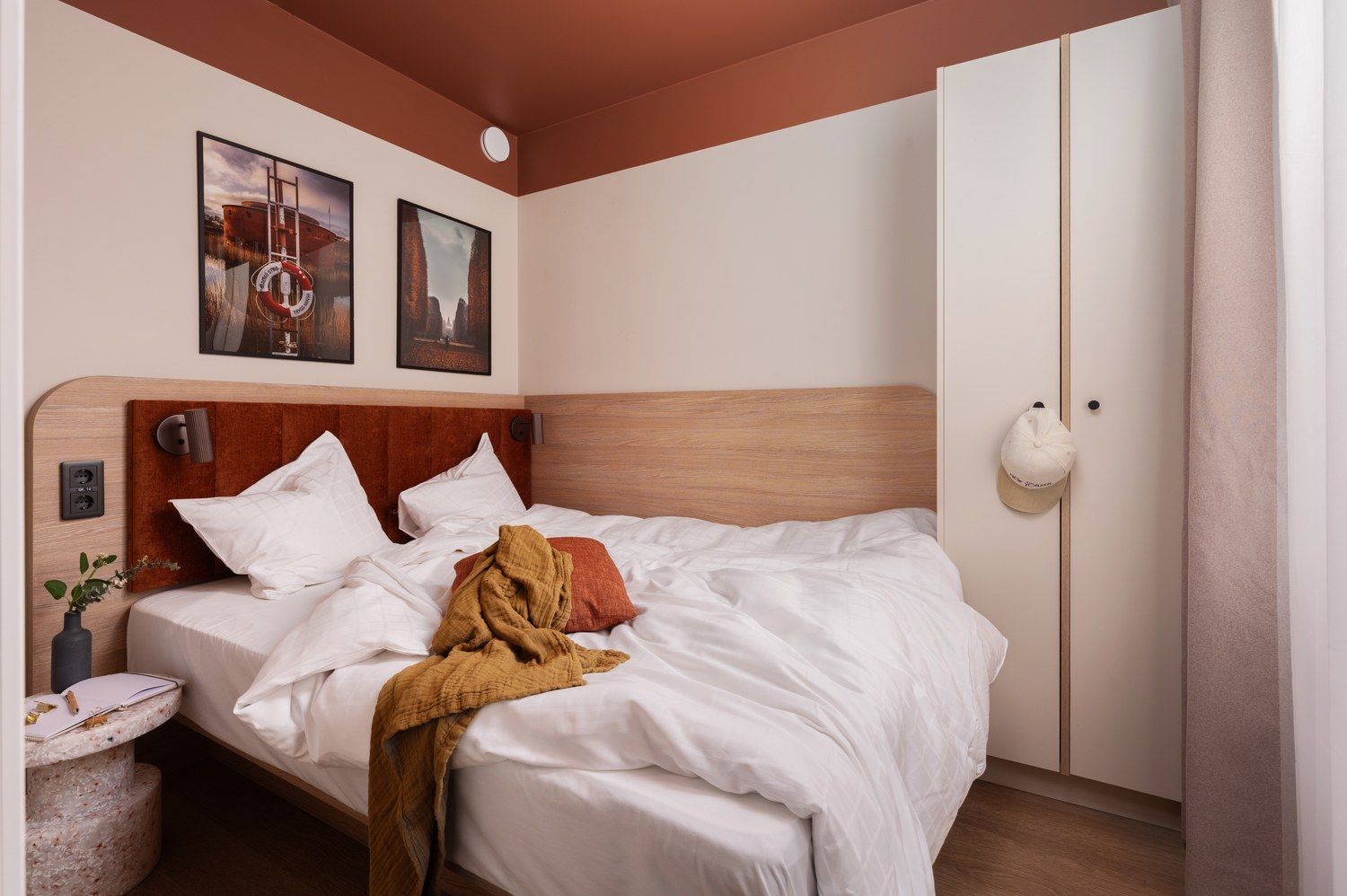 Una cama bien hecha con ropa blanca, cabecera de madera y una manta marrón. Hay dos cuadros enmarcados en la pared sobre la cama, una mesita de noche con un florero y una libreta, y un armario blanco con espejo.