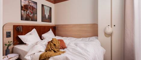 Una cama bien hecha con ropa blanca, cabecera de madera y una manta marrón. Hay dos cuadros enmarcados en la pared sobre la cama, una mesita de noche con un florero y una libreta, y un armario blanco con espejo.