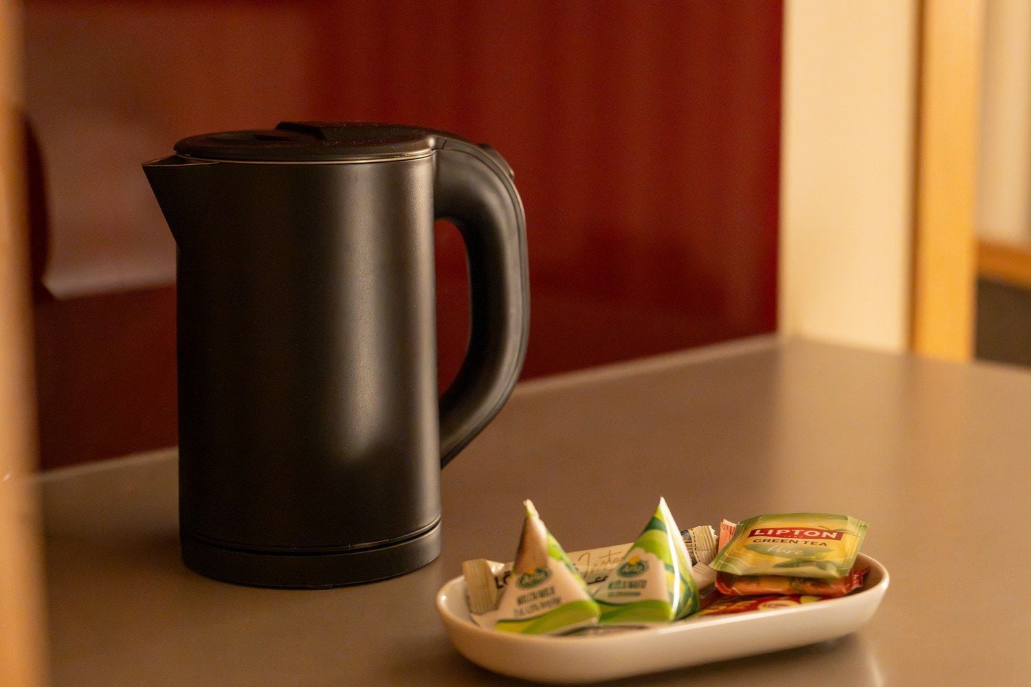 A black electric kettle and a white plate with green and yellow tea packets on a countertop.
