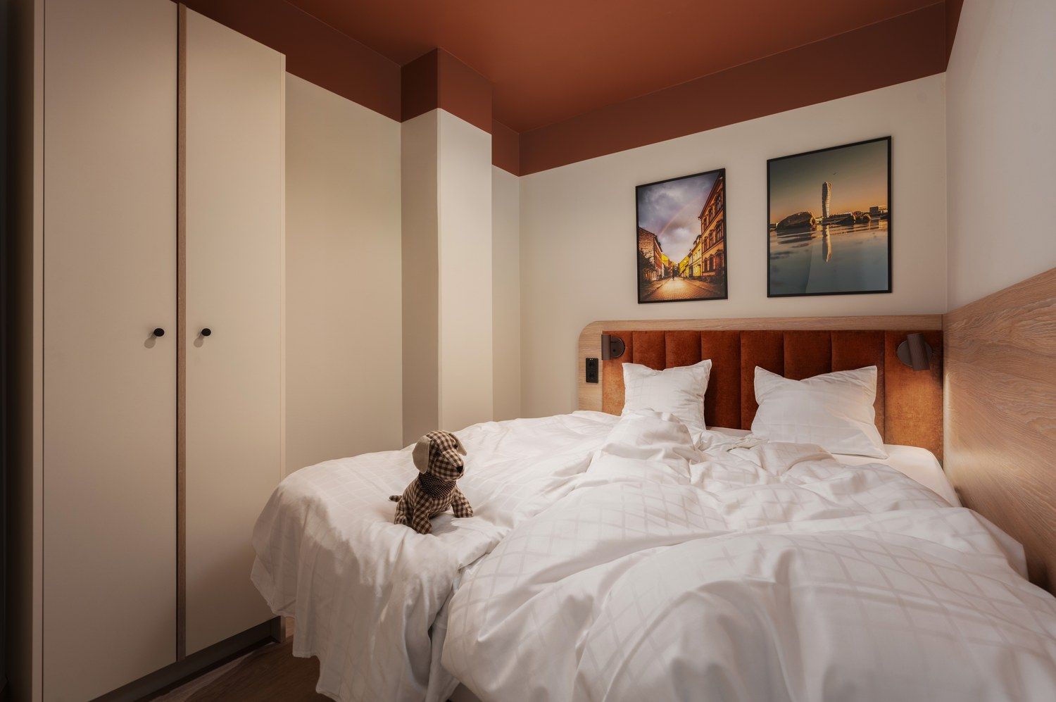 A bedroom with a bed, a headboard, two wall-mounted paintings, and a wardrobe.