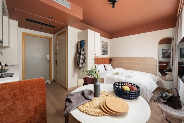 Quarto de hotel com cama, mesa de jantar com cesta de frutas, cadeira e pequena mesa com um vaso.