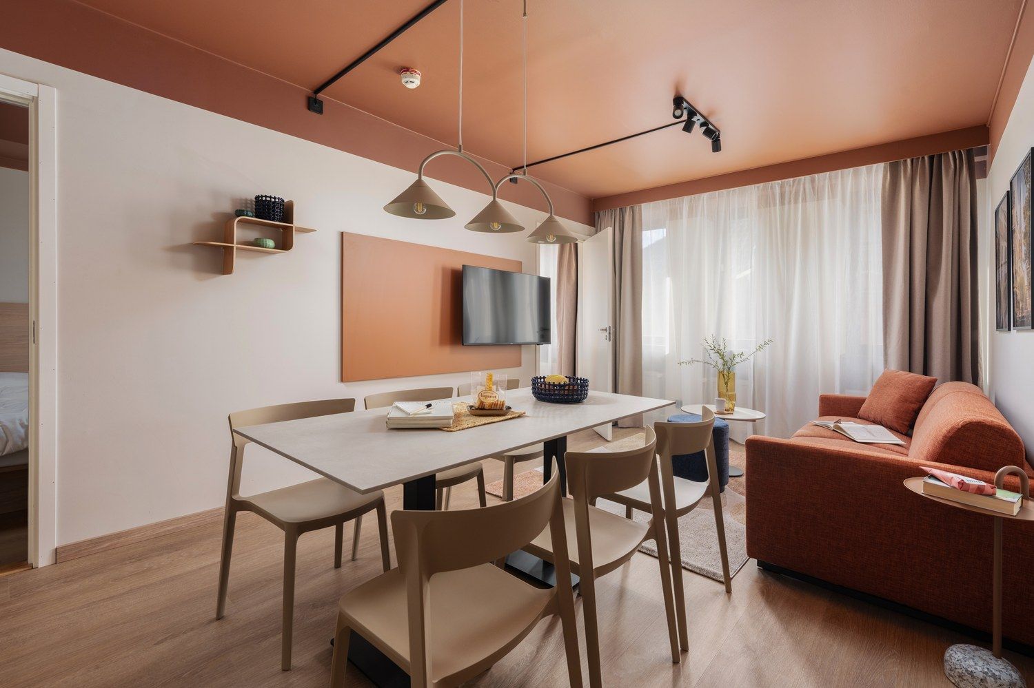 Una sala moderna con un sofá, una mesa de comedor con sillas, un televisor de pantalla plana montado en la pared y una ventana con cortinas.
