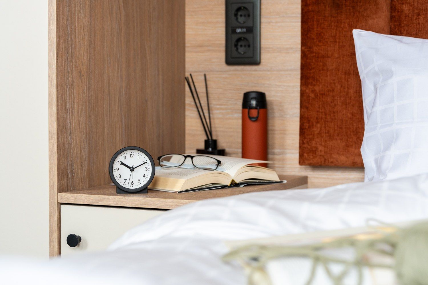 A neatly made bed with white bedding, a bedside table with a clock, an open book, a pair of glasses, and a red candle holder.