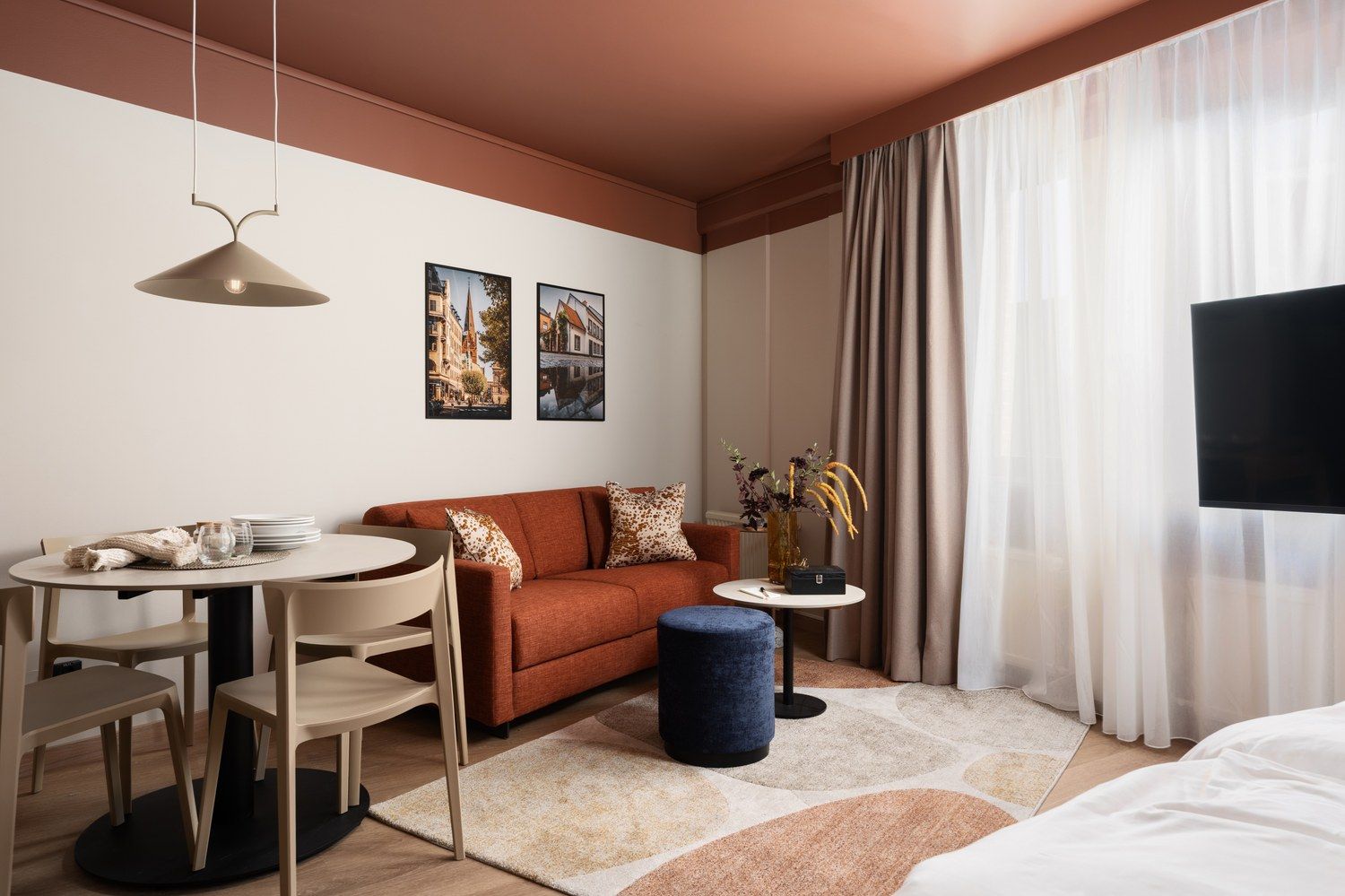 A hotel room with a sofa, a dining table with chairs, a small round table with a vase, and a bed.